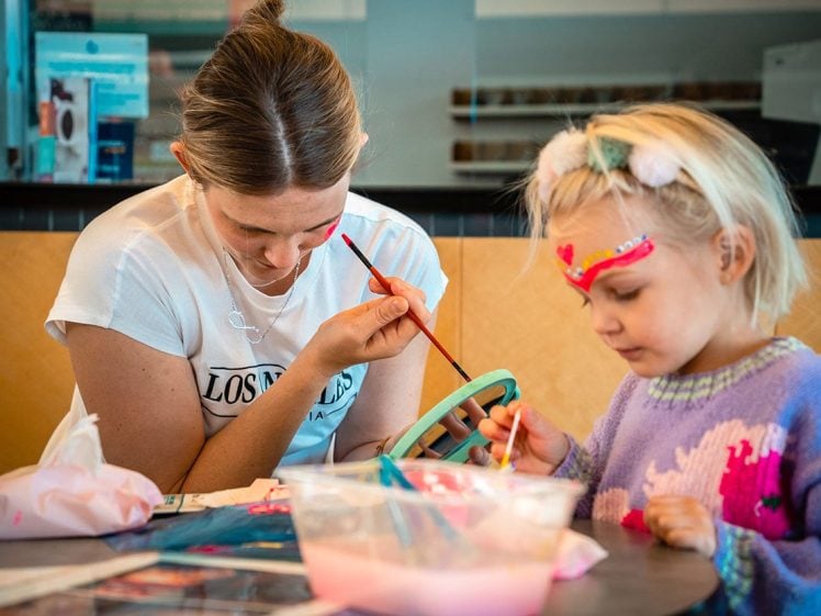 Image of A woman and a young girl are painting together at a table. The girl has a painted heart design on her face and is focused on her artwork, whilst the woman looks down, holding a paintbrush. Art supplies are on the table.