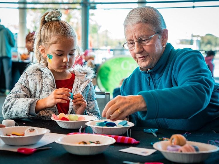 Image of An older woman and a young girl sit at a table, making crafts with colourful felt and thread. They are focused on their work, surrounded by bowls of craft supplies in a bright, indoor setting.
