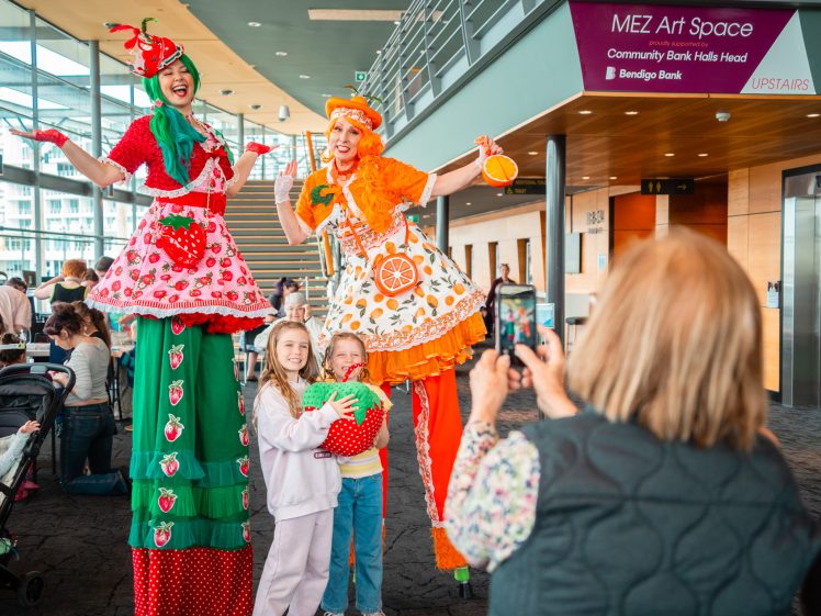 Image of Two performers on stilts in colourful fruit-themed costumes pose with two smiling children holding a giant strawberry as a woman takes their photo indoors near a sign for MEZ Art Space.