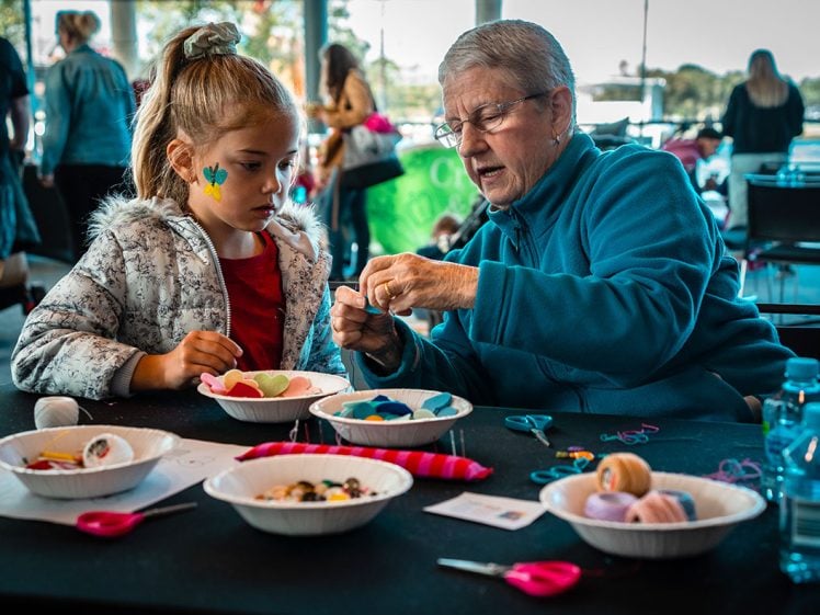 Image of An older woman helps a young girl with a craft project at a table covered with bowls of colourful supplies, scissors, and wool. The girl has a green shamrock painted on her face. Other people are in the background.