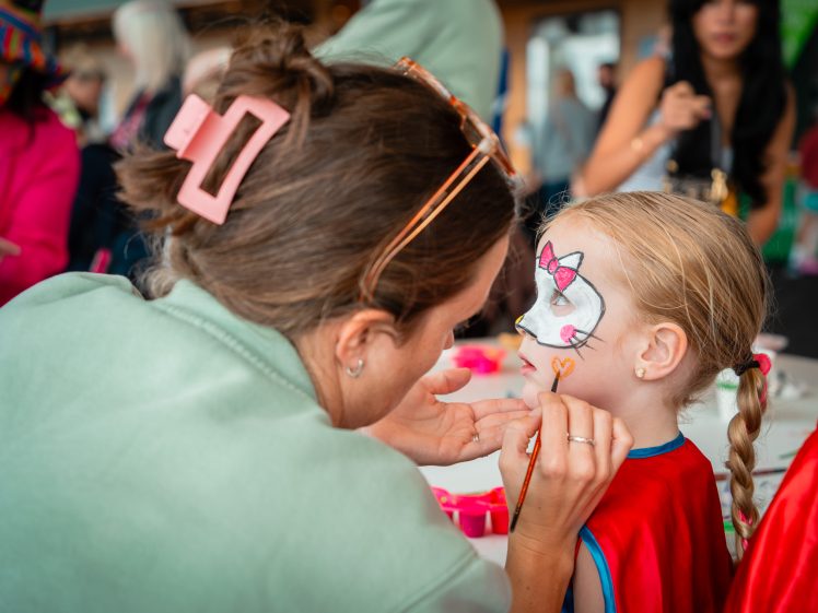 Image of A woman paints a colourful unicorn and heart on a young girl’s face at a busy event. The girl, wearing a red cape, sits still whilst others and vibrant decorations are visible in the background.