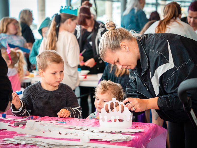 Image of A woman helps two young children with a crown craft at a busy, colourful table during a group art activity. Other children and adults are visible in the background, engaged in similar creative projects.
