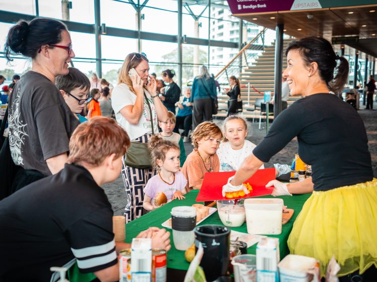 Image of A smiling woman in a yellow skirt demonstrates a cooking activity at a table to a group of children and adults gathered round, watching with interest in a bright indoor space.