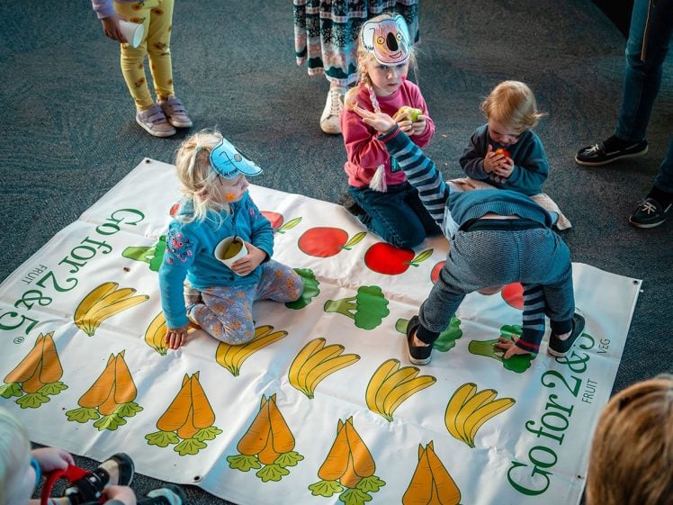 Image of Four children play a fruit-and-vegetable-themed floor game. Two kids wear paper hats, one helps another stand, while others watch. The mat has images of carrots, bananas, apples, and broccoli. Adults' legs are visible nearby.