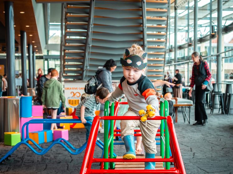 Image of A young child in animal-themed face paint climbs a colourful play structure indoors, surrounded by other children and adults in a bright, modern space with large windows and unique architecture.