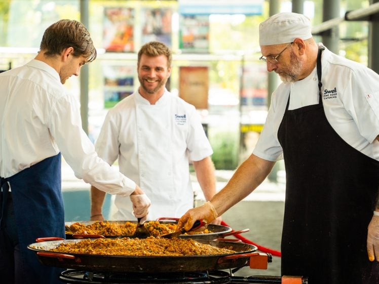 Image of Three chefs in white uniforms and aprons are preparing food together in large pans. Two are serving the food while one smiles in the background. The setting appears to be a modern, well-lit kitchen or event space.