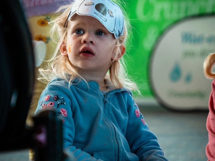 Image of A young child wearing a handmade paper animal mask sits cross-legged on the ground, looking up, holding a cup. The child is dressed in a blue jacket and appears to be at an indoor event or fair.