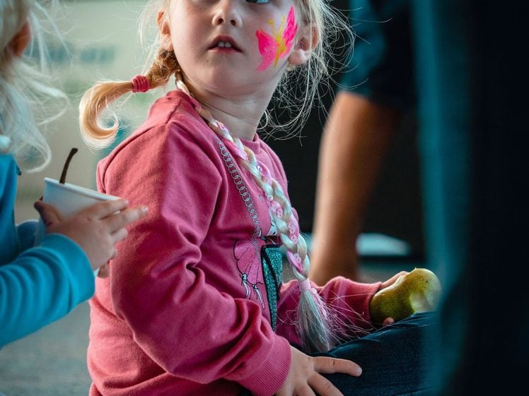 Image of A young girl with a painted butterfly on her cheek and a paper animal mask on her head sits on the floor holding a green apple, looking up. She wears a pink jumper and jeans. Another child sits next to her.