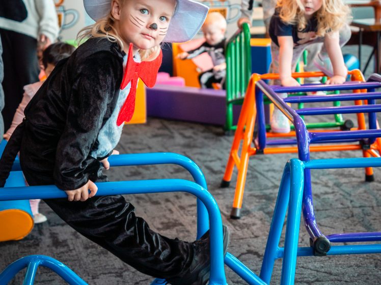 Image of A young child dressed as the Cat in the Hat, with a red and white striped hat, red bow tie, and painted whiskers, sits on blue play equipment indoors. Other children play nearby.