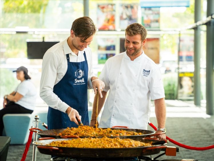 Image of Two men, one in a white chef's jacket and one in a blue apron, stand side by side cooking a large pan of paella indoors. Both are smiling and engaged in preparing the dish together.