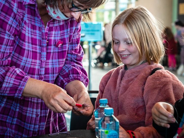 Image of A woman in a checked shirt helps a smiling young girl with a craft project at a table with colourful materials and two water bottles, in a bright indoor setting.