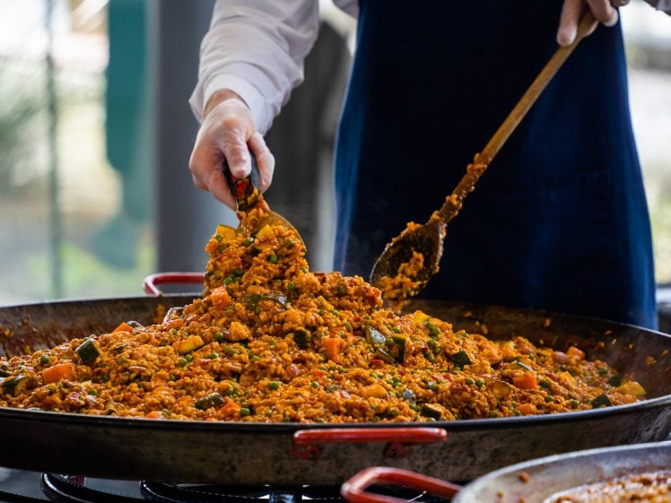 Image of A person in a blue apron serves a large pan of colourful paella using a ladle and spoon, with visible pieces of rice, vegetables, and possibly seafood or meat.