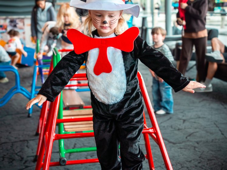 Image of A child dressed as the Cat in the Hat, wearing a tall red-and-white striped hat and a big red bow tie, balances on a wooden bridge at an indoor play area with other children and adults in the background.