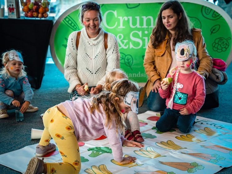 Image of Five young children wearing animal masks play a colourful floor game with fruit and vegetable illustrations, while two women watch and smile. A green Crum & Sip sign is in the background, and a fruit display is to the side.