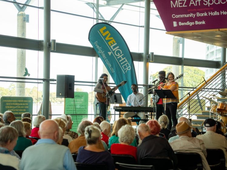 Image of A small band performs on stage in front of a seated audience at an indoor venue with large windows. A LiveLighter banner and musical instruments are visible, and natural light fills the space.