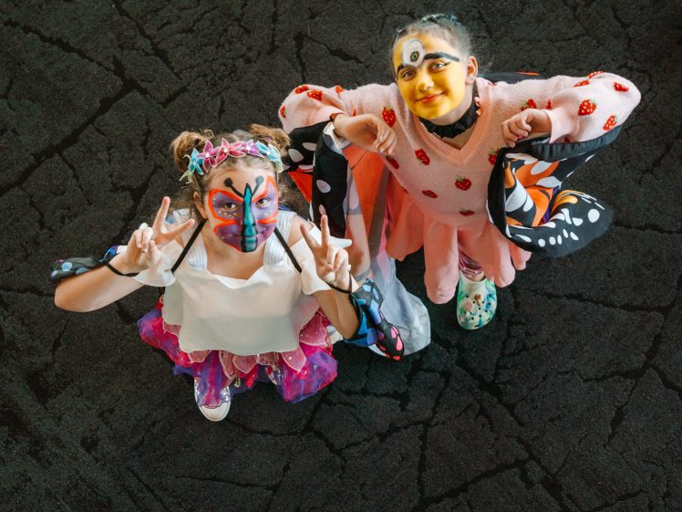 Image of Two children in colourful costumes and face paint stand together, smiling and making peace signs whilst looking up at the camera. The dark cracked floor contrasts with their bright outfits and butterfly wings.