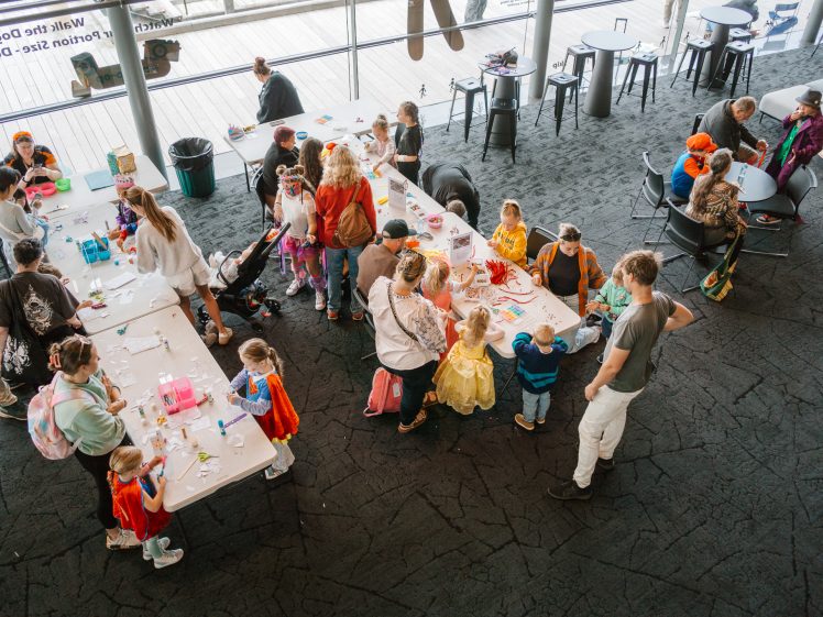 Image of Children and adults, many in fancy dress, gather around tables with craft supplies in a bright indoor space. Some are making crafts, while others watch or chat. The scene is lively and busy.