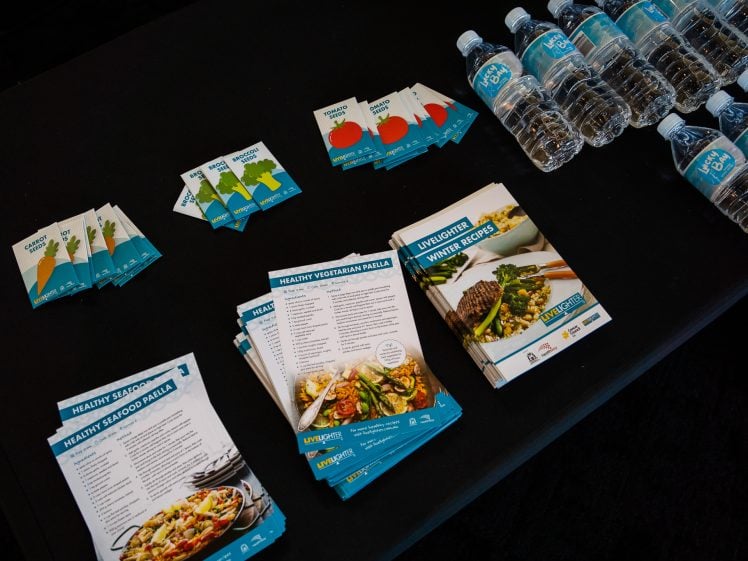 Image of A black table displays bottled water, recipe cards for healthy seafood and vegetarian paella, brochures titled “Live Lighter Winter Recipes,” and small informational cards with vegetable illustrations.