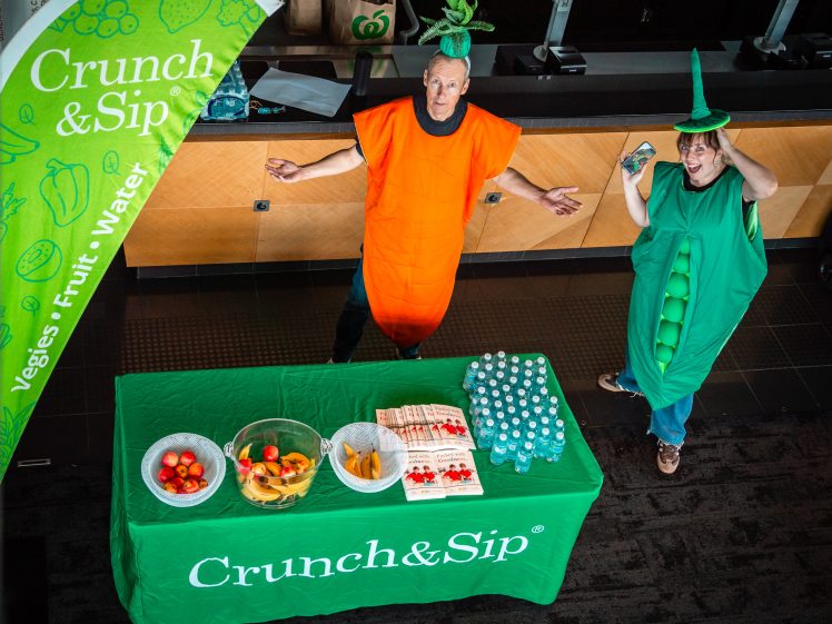 Image of Two people dressed as a carrot and a pea pod stand behind a table with fruit, veg, and water bottles, next to a green Crunch & Sip banner promoting healthy snacks and hydration.