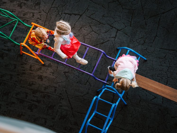 Image of Three young children in colourful clothes play on a climbing frame with ladders, beams, and steps, viewed from above on a dark textured floor.