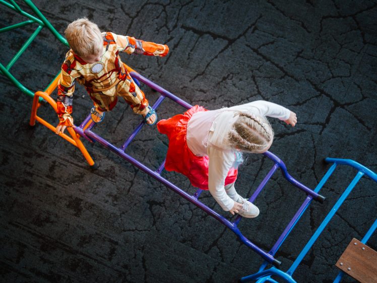 Image of Two young children, one dressed in an Iron Man costume and the other in a white top with a red skirt, walk across colourful metal climbing bars on a dark, textured playground surface, viewed from above.