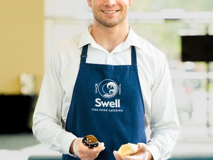 Image of A man wearing a white shirt and a blue apron labelled Swell Fine Food Catering serves food with a ladle and holds a piece of bread, smiling at the camera in a brightly lit indoor setting.
