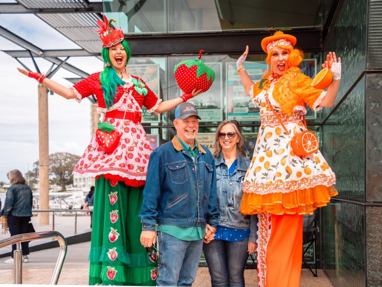 Image of Two people stand smiling, holding hands, between two women on stilts dressed in colourful fruit-themed costumes—one in strawberries and green, the other in oranges and orange hues—outside a modern building.