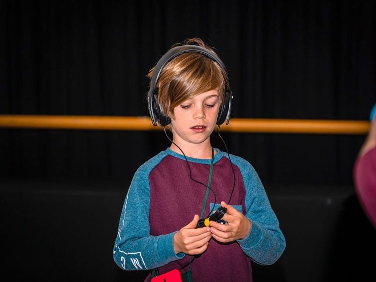 Image of A young boy wearing headphones and a maroon and teal shirt is looking down and holding a small electronic device. The background is dark with a yellow horizontal bar.