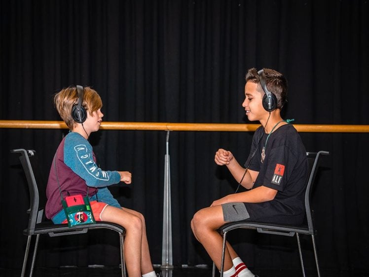 Image of Two boys sit facing each other on chairs, wearing headphones and smiling, against a dark curtain background. One boy wears a blue shirt and red pouch, the other wears a black shirt and grey shorts.