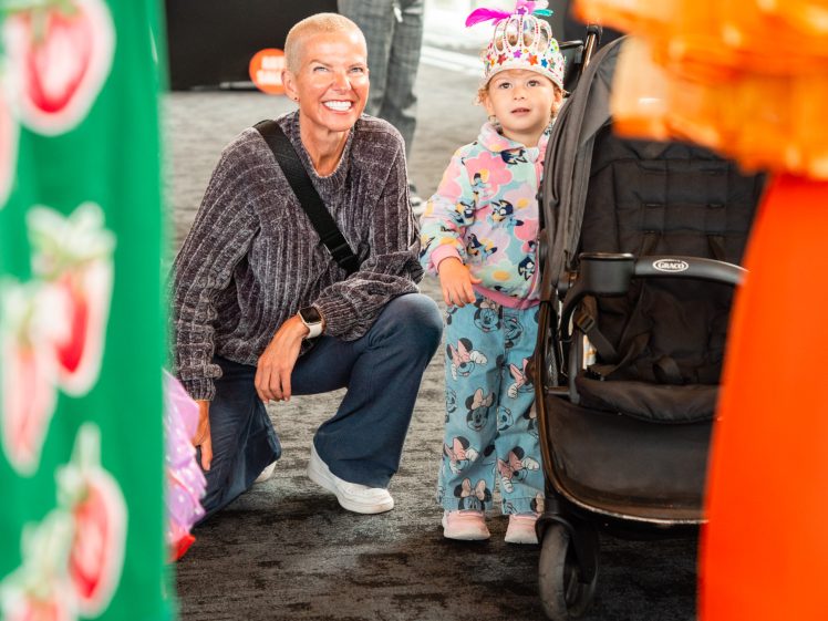 Image of A smiling, bald adult kneels next to a young child in pyjamas and a festive crown, standing by a pushchair indoors. Colourful, out-of-focus costumes frame the image in the foreground.
