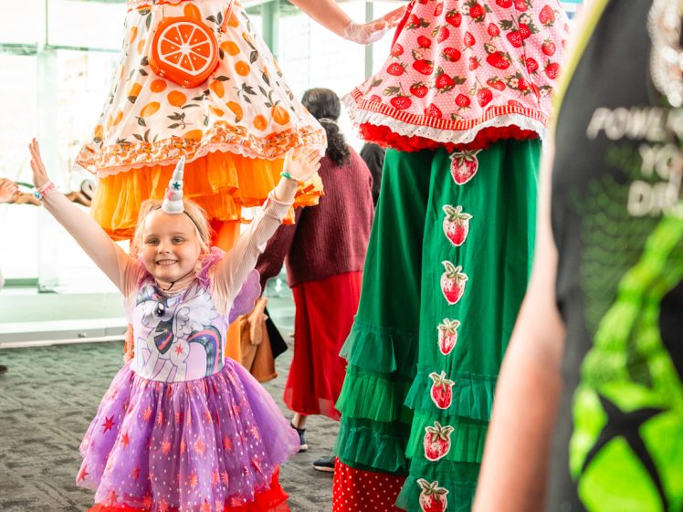 Image of A smiling child in a unicorn costume poses with arms raised beside two women on stilts dressed in colourful fruit-themed outfits, one orange and one strawberry, at an indoor event.