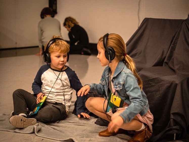 Image of Two young children wearing headphones sit on a grey blanket, talking and interacting. In the background, two other children sit on the floor, also wearing headphones, focussed on their own activities.