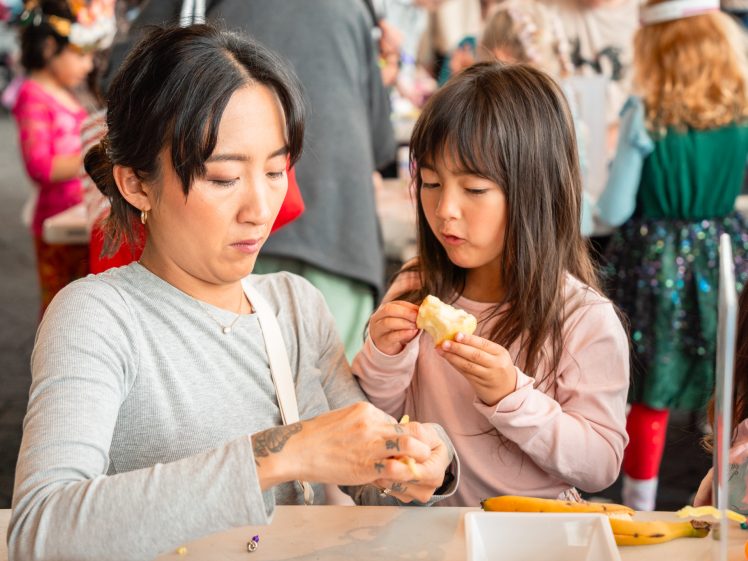 Image of A woman and a young girl sit at a table together. The woman peels a banana whilst the girl, holding part of the banana, watches intently. Other children and adults are visible in the busy background.