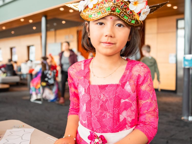 Image of A young girl wearing a vibrant pink dress and an ornate headdress decorated with flowers and gold spikes stands at a table with arts and crafts materials in a bright, modern indoor space.