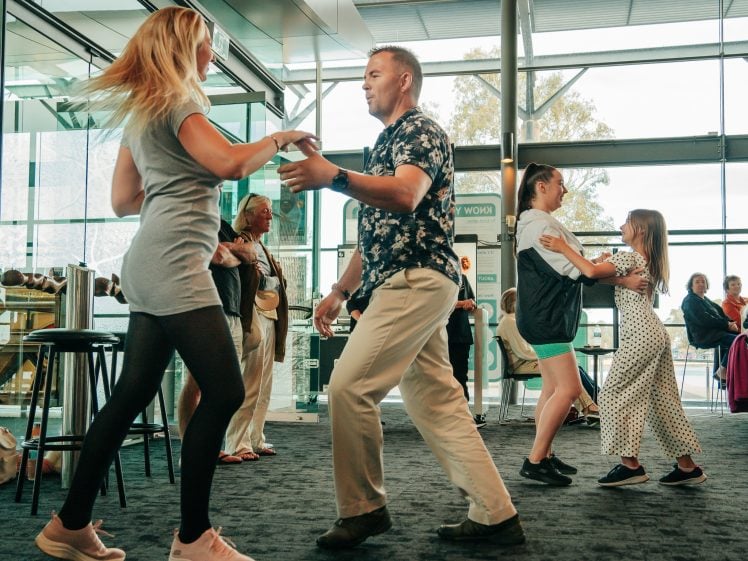 Image of People of various ages dance in pairs inside a modern, sunlit building with large windows. Onlookers stand watching, as two adult pairs and one pair of children practise dance moves on a dark carpeted floor.