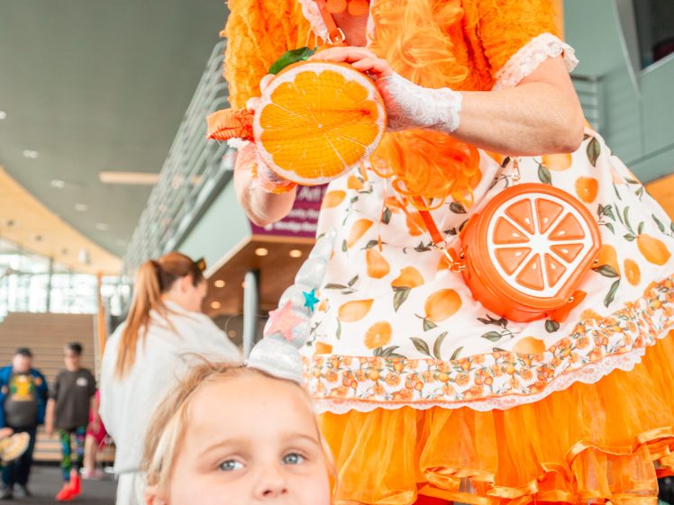 Image of A performer dressed in a vibrant orange-themed costume poses energetically with props resembling orange slices, whilst a young child in a purple costume looks towards the camera indoors at an event.