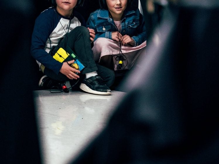 Image of Two children wearing headphones sit on the floor, watching something off-camera. The boy holds a Rubik’s Cube, and the girl sits beside him, both appearing focused. The foreground is partially obscured by dark shapes.