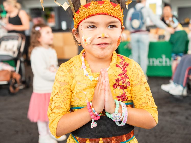 Image of A young girl dressed in colourful traditional attire and a golden headdress smiles with her hands pressed together in a greeting gesture, standing indoors at a lively event with people in the background.