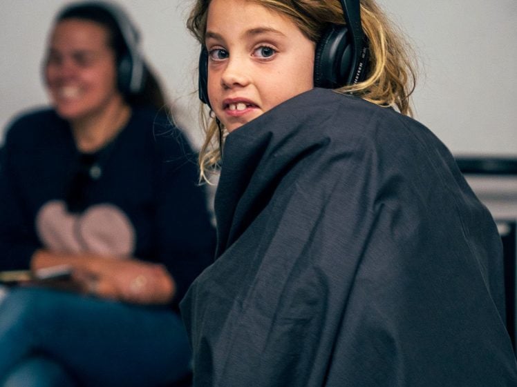 Image of A young girl with blonde hair wears headphones and is wrapped in a dark blanket. She looks towards the camera. In the background, an adult woman sits, smiling and also wearing headphones.