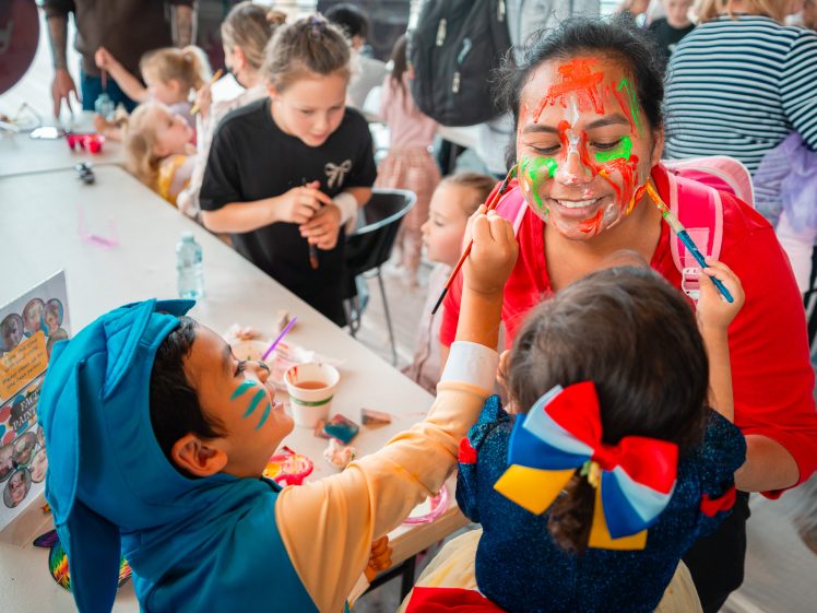 Image of Two children in fancy dress paint a woman’s face with bright colours at a crowded arts and crafts table, surrounded by other kids and adults engaged in creative activities.