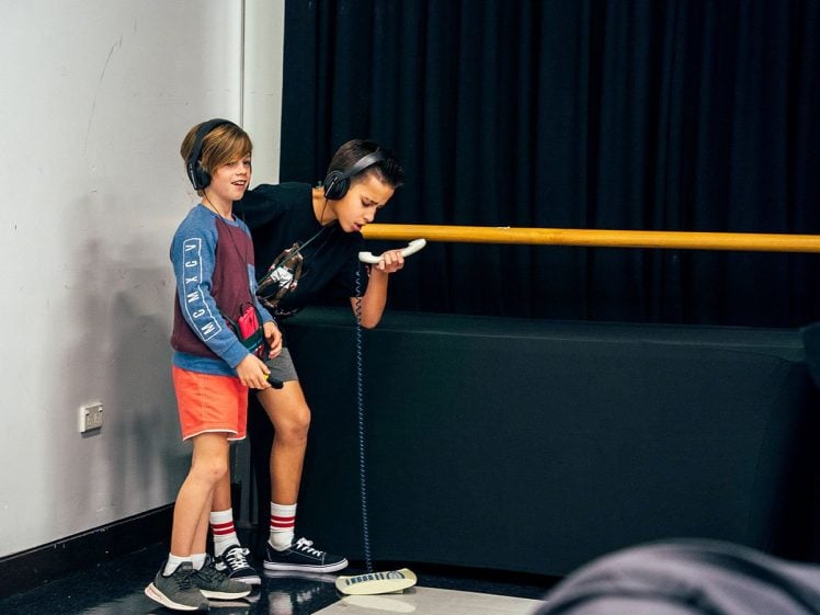 Image of Two boys wearing headphones stand near a wooden rail. One boy holds a microphone and leans forward to speak or sing, whilst the other smiles and looks on. There is a black curtain in the background.