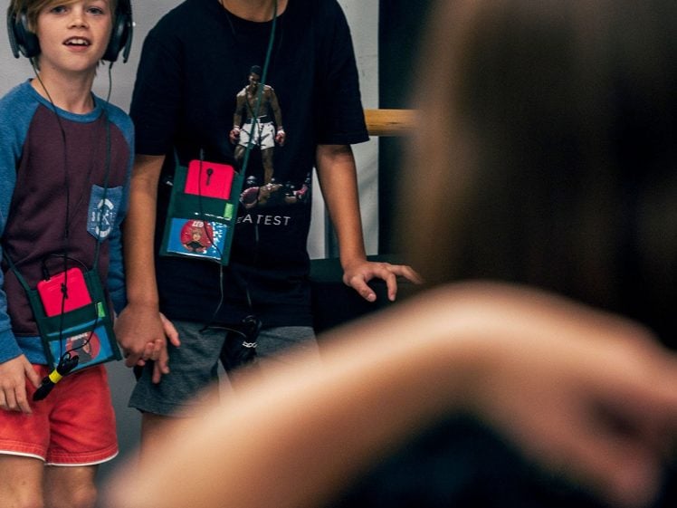 Image of Two boys wearing headphones and colourful badges stand side by side, watching a person in the foreground with their arm raised. The boys appear engaged and interested in what’s happening.