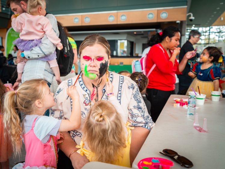Image of Two young girls in fancy dress paint a seated woman’s face with bright colours at a busy indoor event, while other children and adults take part in similar activities in the background. A table with art materials is in the foreground.