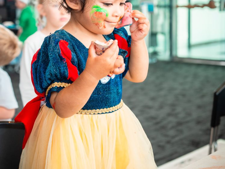 Image of A young child in a Snow White fancy dress paints her face with colourful face paint, smiling as she looks in a small mirror on the table in front of her. Art supplies and drawings are scattered across the table.