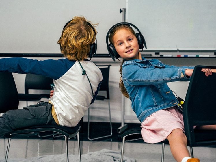 Image of Two children sit back-to-back on chairs, wearing headphones and smiling. They are in a classroom with a whiteboard and empty chairs in the background. The girl faces the camera whilst the boy looks away.