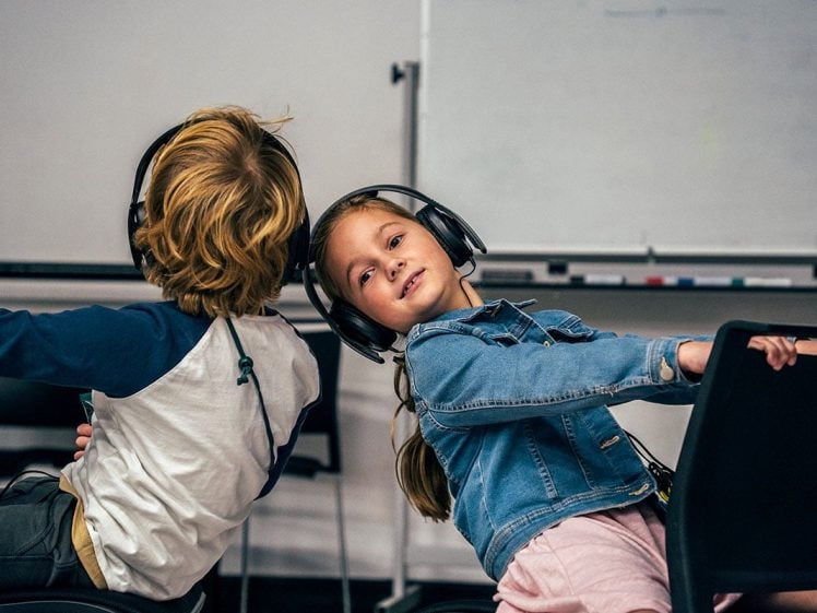 Image of Two children wearing headphones sit back-to-back on chairs, turning towards opposite sides. The girl looks at the camera and smiles, whilst the boy faces away. A whiteboard is visible in the background.