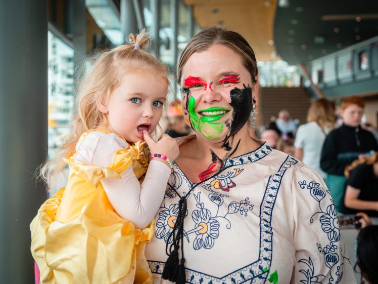 Image of A woman with colourful face paint smiles whilst holding a young girl in a yellow princess dress. They are indoors, surrounded by people in a busy, bright setting.