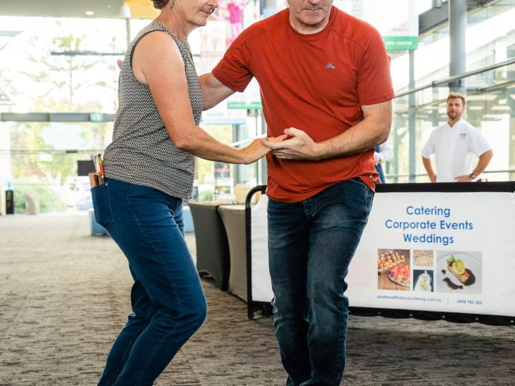 Image of A woman and a man dance together indoors on a carpeted floor, holding hands. They are casually dressed, and there is a sign in the background advertising catering for corporate events and weddings.