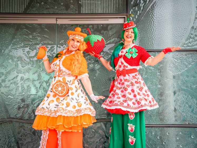 Image of Two women in colourful fruit-themed costumes pose and smile. One wears orange attire with orange slice patterns, and the other wears red-and-green clothes with strawberry motifs. Both stand before a textured glass wall.
