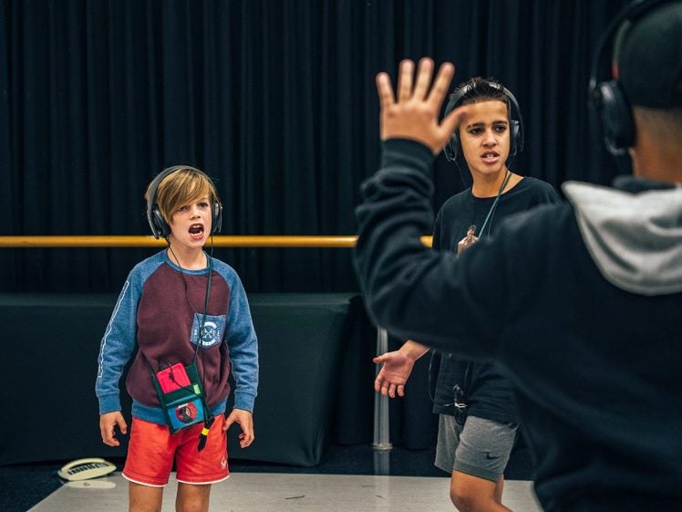 Image of Two children wearing headphones stand facing an instructor who raises a hand. The children appear focused and engaged, possibly singing or reacting to instructions in a studio with a black curtain in the background.